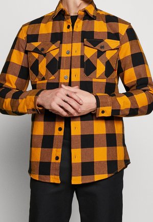 Hombre con camisa de manga larga a cuadros amarillos y negros con bolsillos en el pecho abotonados, de pie con las manos entrelazadas al frente.