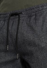 Šedé sportovní kalhoty s elastickým páskem na šňůrku, které mají vzor rybí kost a kovové koncovky na šňůrce.