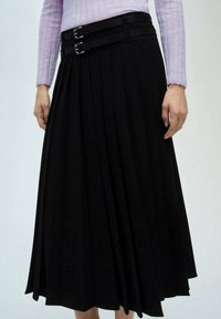 Zwarte plissee rok met twee horizontale stoffen bandjes op de taille, voorzien van zilveren gespen. De stof lijkt glad en ondoorzichtig.