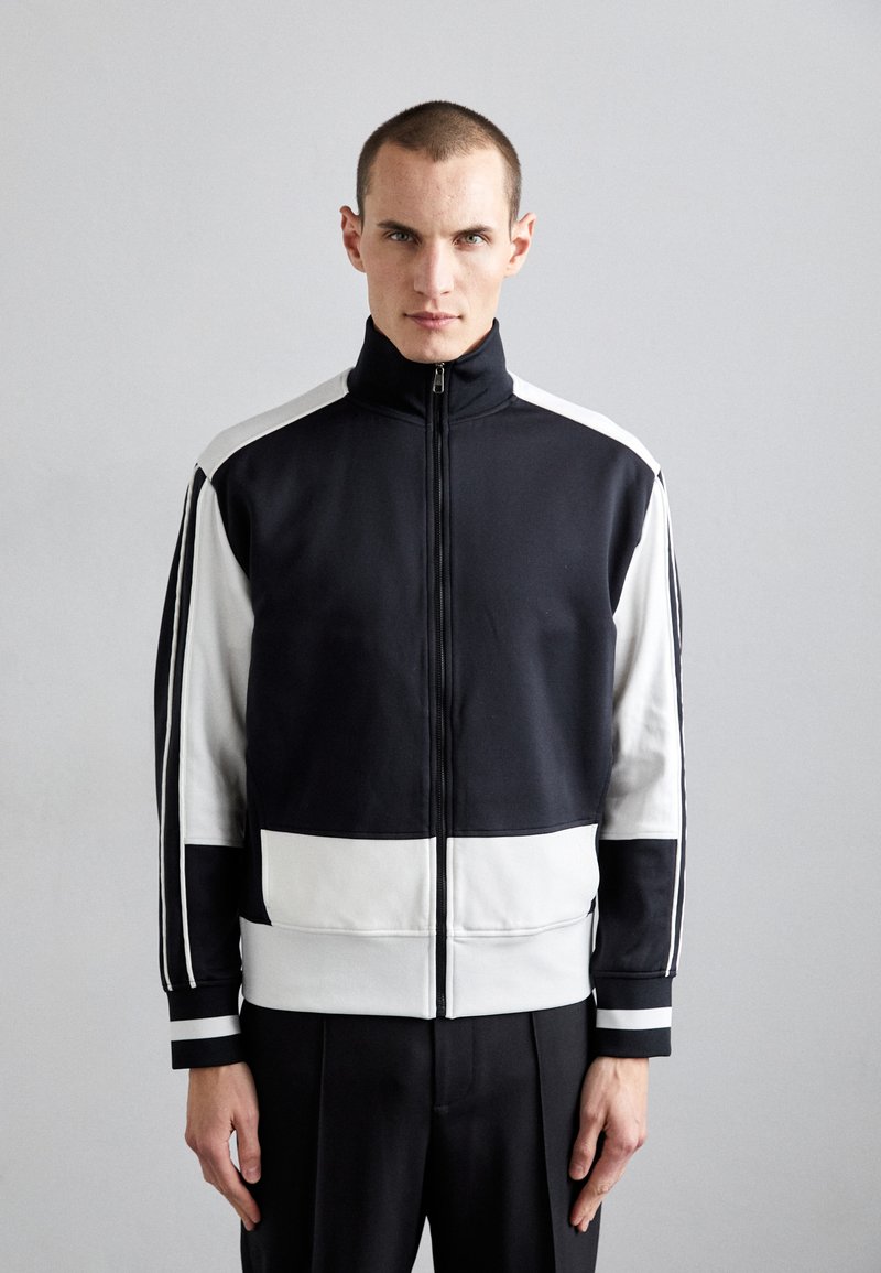 Black and white zip-up jacket with a high collar, featuring bold color blocks and contrasting stripes on the sleeves and cuffs. Smooth fabric texture.