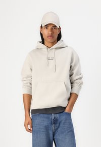 Felpa grigia chiaro con tasca frontale e cappuccio con coulisse, abbinata a jeans blu. La felpa presenta un piccolo logo nero sul petto.