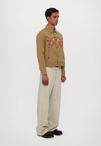 Tan button-up jacket with floral embroidery, black stitching details; paired with light gray wide-leg trousers and black footwear.