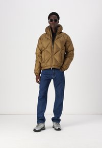Giacca puffer marrone con cuciture a forma di diamante, chiusura con zip e cappuccio regolabile, indossata con jeans scuri e sneakers grigie.