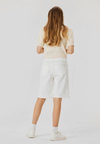 Kvinna med långt brunt hår som bär en krämfärgad kortärmad tröja, vita knälånga shorts, vita strumpor och vita sneakers, stående framför en vit bakgrund.