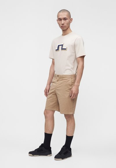 Homme debout portant un short beige, un t-shirt crème avec un logo géométrique, des chaussettes noires et des baskets noires sur un fond uni.