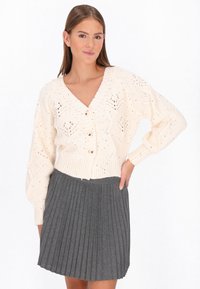 Femeie îmbrăcată cu un pulover crem tricotat, cu nasturi și model de frunze, și fustă plisată gri, stând cu mâna pe șold, pe un fundal alb.