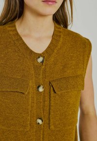 Gilet lavorato a maglia giallo senape con scollo a V, due tasche frontali e bottoni marroni. Presenta un tessuto texturizzato e bordi a coste.