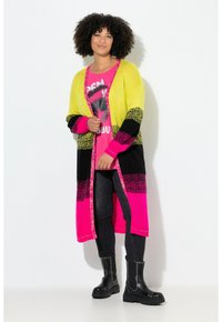 Lange Strickjacke in leuchtendem Gelb, Pink und Schwarz mit einem gemusterten Design. Getragen über einem pinkfarbenen Grafik-T-Shirt und kombiniert mit schwarzen Knöchelstiefeln.