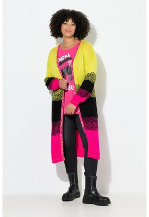 Long cardigan en jaune vif, rose et noir avec un motif. Porté sur un T-shirt graphique rose et associé à des bottines noires.