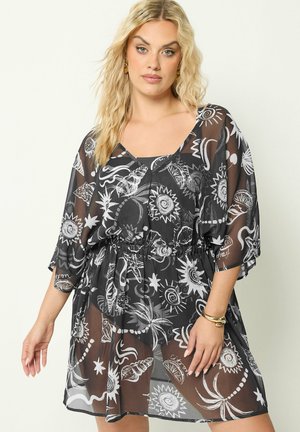 Femme aux longs cheveux blonds portant une robe noire transparente avec des motifs abstraits blancs de soleil et de coquillages, accessoirisée avec des boucles d'oreilles et des bracelets en or.