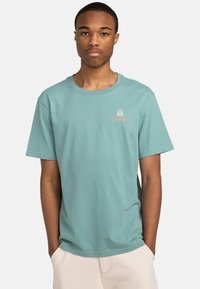Camiseta de algodón azul claro con cuello redondo, mangas cortas y un pequeño logo en blanco y naranja de "Element" que presenta gráficos de árboles en el pecho.