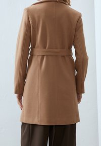 Manteau en laine marron avec taille ceinturée, coupe cintrée, manches longues et col cranté. Texture lisse avec détails de coutures visibles.