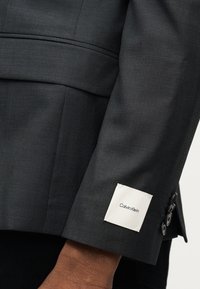 Μανίκι σκούρου γκρίζου κοστουμιού με τρία μαύρα κουμπιά και μια λευκή ορθογώνια ετικέτα Calvin Klein ραμμένη κοντά στη μανσέτα πάνω σε σκούροχρωμο χέρι.