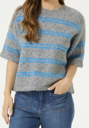 Femme portant un pull en maille gris avec des rayures horizontales bleues et un jean bleu, avec une main dans la poche et les cheveux lâchés sur l'épaule.