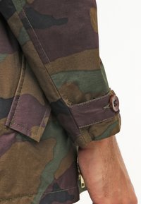 Manžeta rukávu maskovací bundy s detailem knoflíku, kterou nosí osoba s viditelnými chlupy na rukou a světlou hnědou pletí.