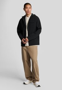 Sort letvægtsjakke med hætte, lynlås foran og brystlomme, båret over en cremefarvet polo, parret med beige bukser og hvide sneakers.