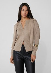 Beige Strickjacke mit rundem Halsausschnitt, Knopfverschluss und schmalen Ärmeln. Kombiniert mit glänzenden schwarzen Lederhosen.