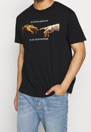 Czarny t-shirt z grafiką dwóch prawie dotykających się dłoni i napisem „ZRÓWNOWAŻONY ROZWÓJ JEST W NASZYCH RĘKACH” noszony przez osobę w jasnoniebieskich dżinsach.