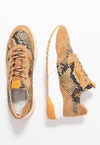 Baskets avec une tige en daim marron clair, des détails en imprimé serpent jaune et noir, un col rembourré, et une semelle en caoutchouc blanche et orange.