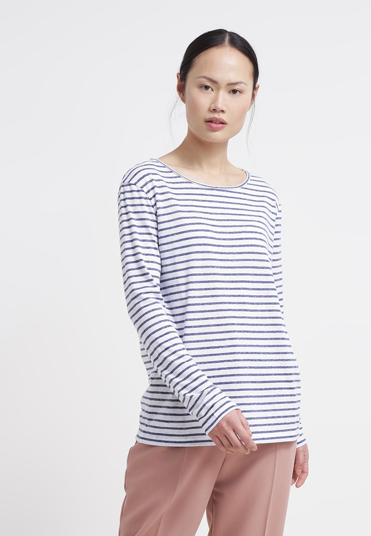 Junge Frau mit dunklem Haar trägt ein weißes und navy gestreiftes Langarmshirt und hellbraune Hose vor einem schlichten Hintergrund.