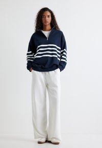 Jersey marino con acentos de rayas blancas, cuello con media cremallera, corte relajado, combinado con pantalones blancos de pierna ancha y calzado casual.