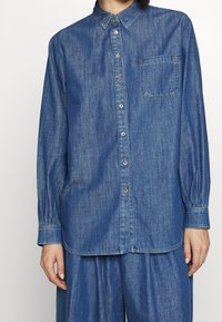 Nainen pukeutuneena siniseen farkkupaitaan, jossa on rintatasku, ja samaan sarjaan kuuluviin väljälahkeisiin farkkuihin, seisoo rento asento kädet vapaasti.