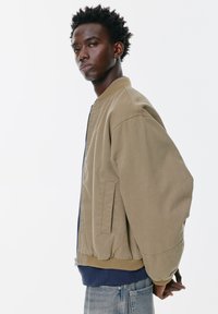 Giubbotto bomber kaki con colletto e polsini a coste, dotato di zip frontale, tasche laterali e tessuto testurizzato. Indossato sopra una camicia blu navy.