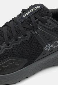 Schwarzer Sportschuh mit einem Obermaterial aus Mesh, strukturierten Akzenten und einer Gummisohle. Die Schnürsenkel sind schwarz, mit einem grauen Logo auf der Zunge.