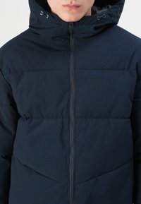 Navy puffer jacket with a zip front, a hood, and a textured fabric. Features horizontal seams and a chevron pattern on the chest.
