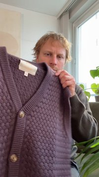 Purpurinis megztas kardiganas su tekstūruotu raštu, gilia V formos iškirpte ir dviem auksiniais mygtukais priekyje. Minkšta medžiaga; ribuotos rankogaliai ir apačia.