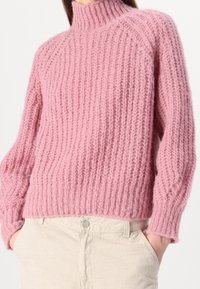 Rosa gestrickter Pullover mit hohem Kragen, gerippter Textur und langen Ärmeln; weiches, flauschiges Material; getragen über hellbeigen Hosen.