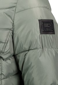 Olive green puffer jacket with a shiny finish, featuring quilted sections, a textured black label with embossed text, and snap hardware accents.