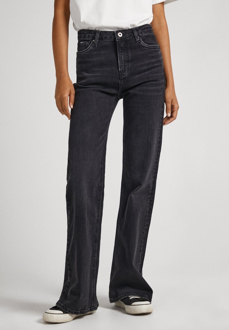 Personne portant un jean noir taille haute à jambes droites et des baskets noir et blanc, debout devant un fond clair uni.