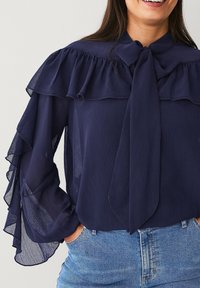 Kvinde iført en gennemsigtig marineblå bluse med volanger på ærmerne og krave, der er bundet i en sløjfe, sammen med lyseblå jeans.