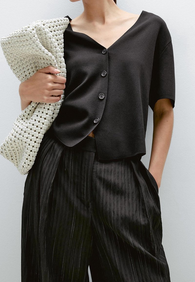 Femme portant un haut noir à manches courtes boutonné et un pantalon large à fines rayures noires, tenant une grande pochette blanche tissée.