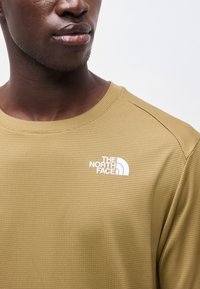 Κοντινό πλάνο ενός ατόμου που φοράει μια υφασμάτινη χακί πράσινη μπλούζα με το λογότυπο The North Face στο στήθος, στραμμένο ελαφρώς αριστερά.