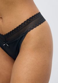 Μαύρο δαντελένιο στρινγκ με κυματιστή άκρη και λεία υφασμάτινη επιφάνεια. Μικρό φιόγκος στη μέση και ορατή ετικέτα με το λογότυπο.