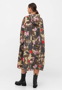 Długi, kapturowy płaszcz przeciwdeszczowy wykonany z wodoodpornego materiału, w brązowym kamuflażu z różowymi i czerwonymi kwiatowymi akcentami. Widoczne czarne botki za kostkę.