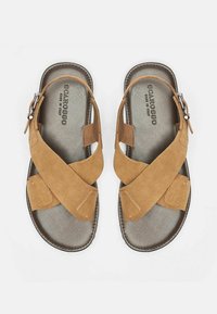 Smeđe suède sandale s križnim trakama i podesivim bočnim kopčanjem. Teksturirana unutrašnjost i glatka potplat. Ime brenda otisnuto unutar.