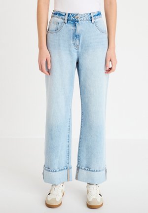 Jeans a gamba larga azzurri con risvolti arrotolati, indossati con sneakers bianche e una maglietta bianca, mostrati dalla vita ai piedi.