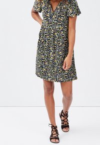 Robe florale noire avec des motifs jaunes et verts, à manches courtes, décolleté en V et taille froncée. Assortie à des sandales noires à talons.