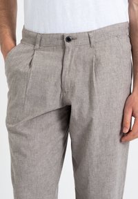 Mężczyzna ubraný w jasnobrązowe lniane spodnie z zapięciem na guzik i zamek błyskawiczny, z bocznymi kieszeniami oraz biała koszulę schowaną w spodnie.