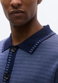 Πολύχρωμο πόλο μπλούζας σε ναυτικό και μπλε με σχέδιο υφασμένης πλέξης, κλείσιμο με  κουμπιά και αντίθετο μονόχρωμο γιακά με γεωμετρικό διακοσμητικό στοιχείο.