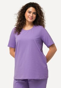 T-shirt viola a maniche corte in tessuto morbido, con scollatura rotonda e vestibilità rilassata. Indossato con pantaloni viola coordinati.