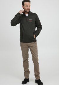 Veste en polaire grise foncée zippée avec un col haut et un logo sur la poitrine. Comprend des accents de fermeture éclair rouges et deux poches latérales.