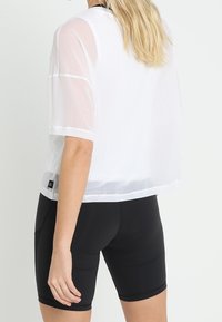 Vit, transparent kortärmad topp med rundad halsringning, lagerdesign och en logotags, kombinerad med svarta åtsittande shorts.