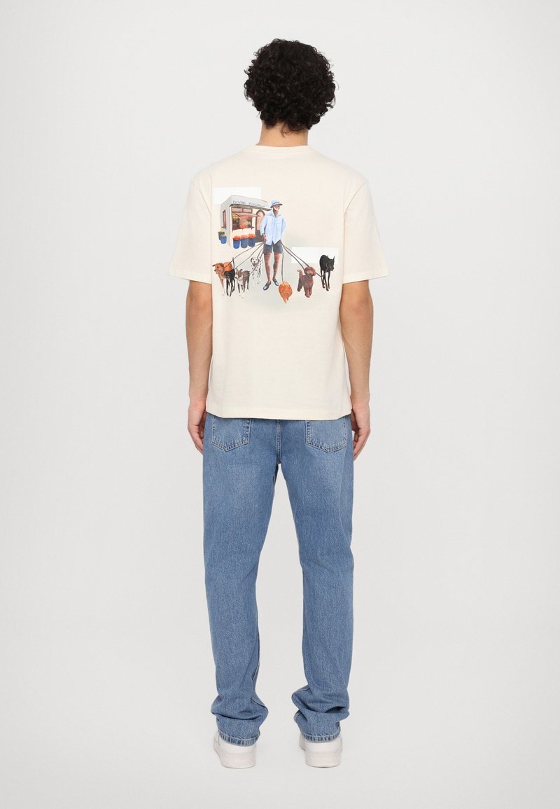 Kremfarget bomull t-skjorte med et fargerikt grafisk trykk av en person som går flere hunder foran en matvogn. Korte ermer og avslappet passform.