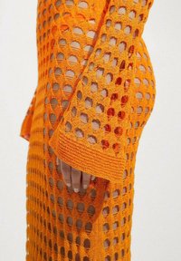 Main portant un tissu en crochet orange translucide avec de grands trous en forme de diamant, montrant la peau en dessous.