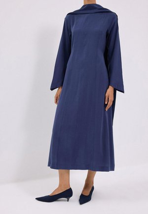 Femme portant une robe bleu marine à manches longues, longueur cheville, avec un large col et des chaussures pointues à talons bas assorties.