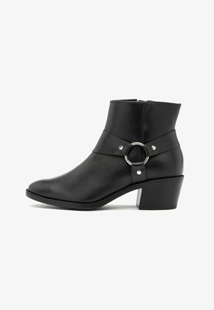 Bottine en cuir noir avec un petit talon carré, une sangle décorative et un détail en anneau argenté sur le côté, sur fond blanc.